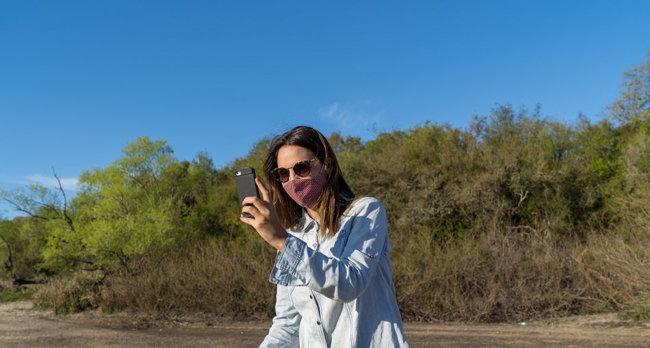 Woman wearing mask using smart phone standing against trees