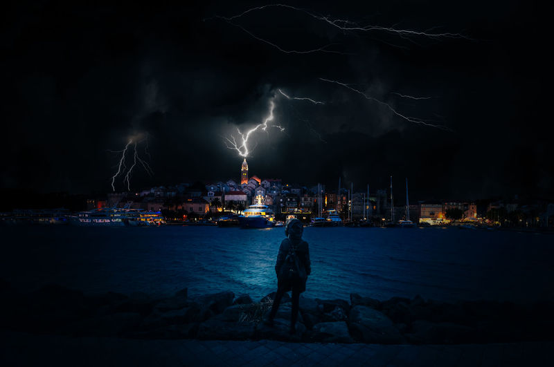 Panoramic view of illuminated city by sea against thunder sky at night