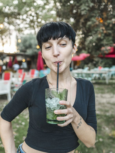 Content female drinking refreshing cocktail with ice cubes and mint leaves while standing in summer park