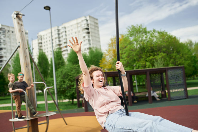 Positive female riding swing rope while laughing and having fun on playground during summer weekend