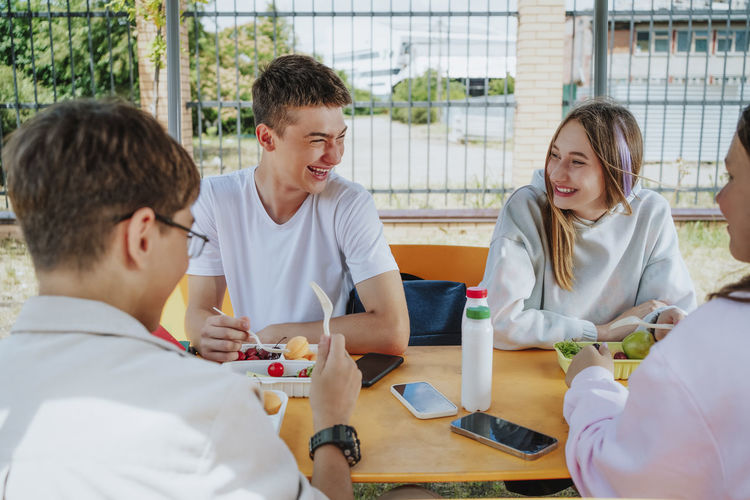 Happy teenage friends having lunch sitting at table in schoolyard