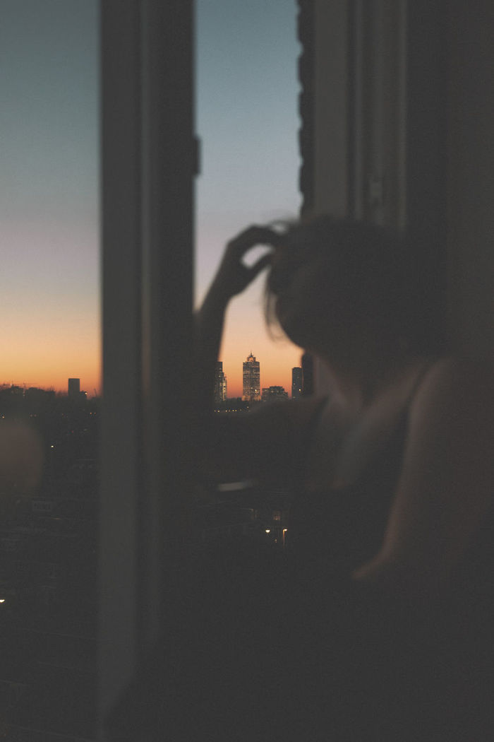 Woman looking away while standing by window at sunset