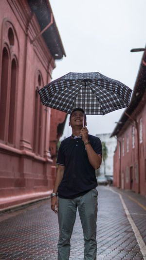 Young man holding umbrella standing on street amidst buildings