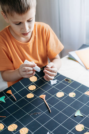 Cute child creating halloween-themed board game. boy gluing paper-cut game elements on playing field