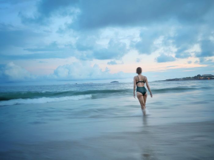 Rear view of bikini woman standing at beach against cloudy sky