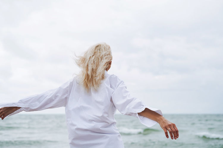 Portrait of elegant blonde woman in white shirt on sand beach at storm sea at windy weater