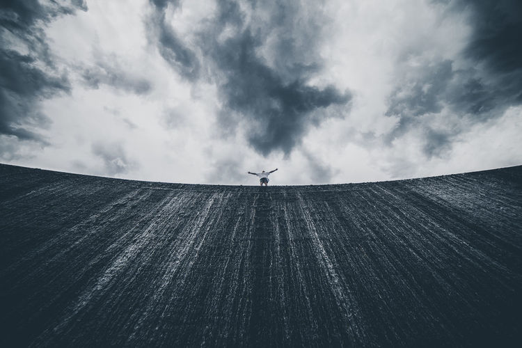 Low angle view of man with arms outstretched standing on wall against cloudy sky