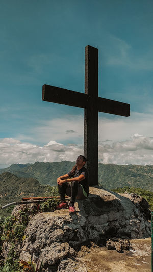Man sitting by cross against sky