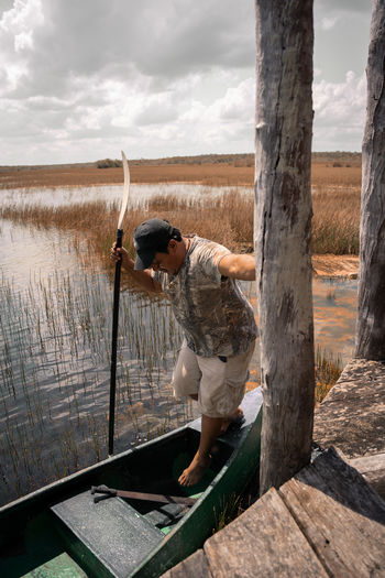 Hispanic rustic male with paddle standing in shabby boat floating on rover near wooden quay in countryside