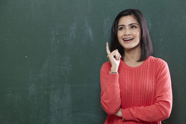 Happy female teacher pointing while standing by blackboard
