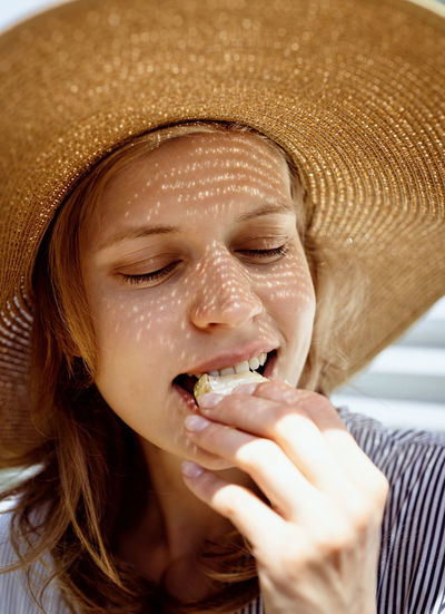 Woman in hat eating grilled vegetables outdoors, eyes closed. light and shadow pattern on the face