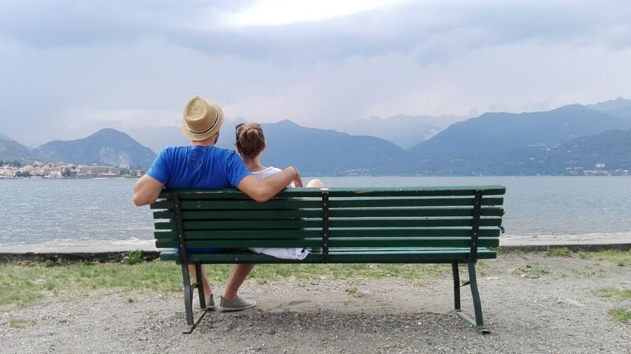 Rear view of man and woman sitting on bench by lake against sky