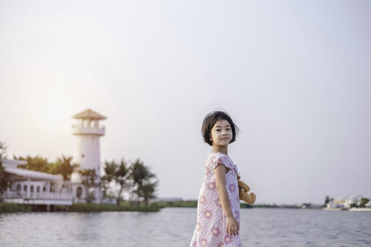 Girl holding teddy bear while standing against lake and sky