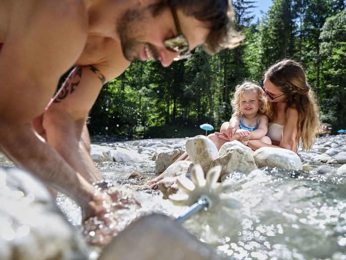 Father fixing water wheel in a mountain stream with mother and daughter in background