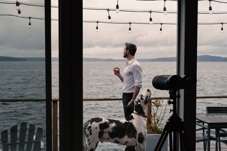 Man holding drink in glass with dog standing by railing and sea against sky