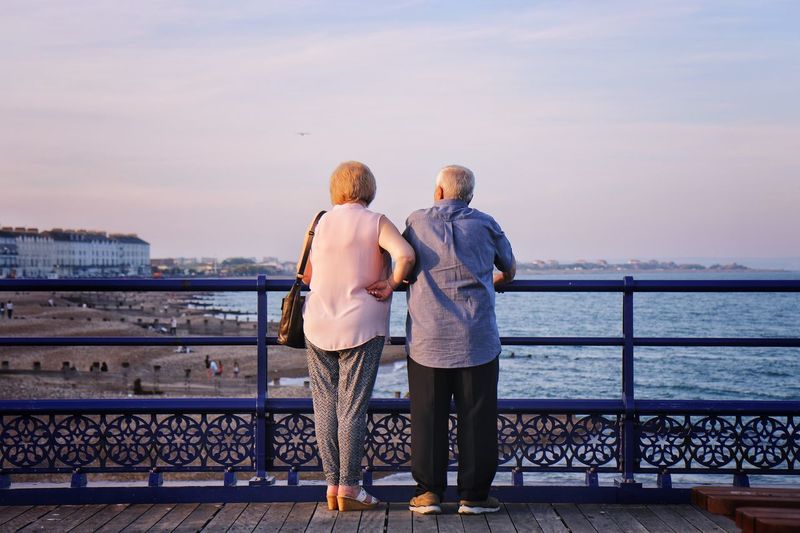 Rear view of couple looking at sea while standing by railing against sky during sunset