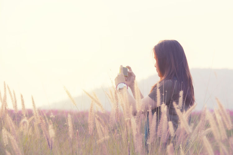 Side view of woman photographing while standing by plants against sky