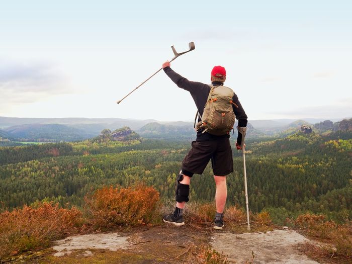 Wounded tourist with crutch above head achieved peak. hiker with backpack and knee brace feature. 