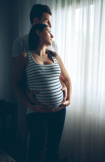 Pregnant woman standing with man against window at home