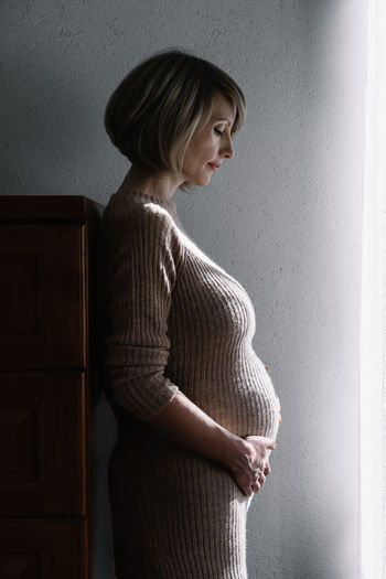 Pregnant woman looking away while standing against wall at home