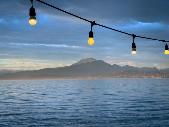 Illuminated light bulbs hanging over sea against sky