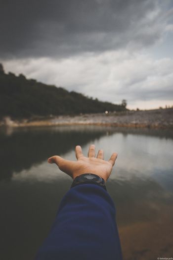 Cropped hand over lake against cloudy sky