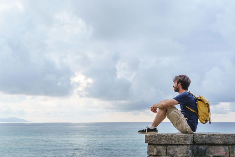Rear view of man sitting on rock by sea against sky
