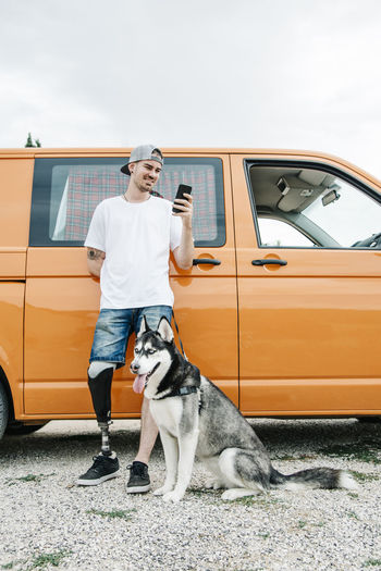 Young man with dog wearing leg prosthesis and using cell phone at camper van
