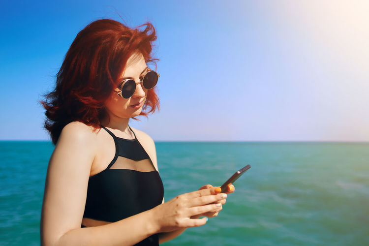 Young woman using smart phone by sea against sky
