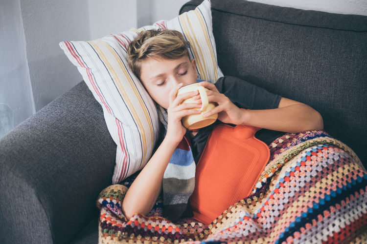 High angle view of ill boy having drink while lying on sofa at home
