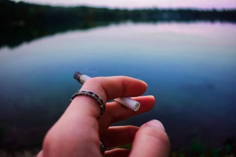 Cropped hand of woman holding cigarette by lake during sunset