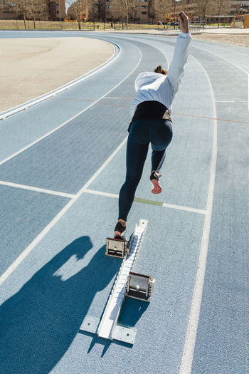 Back view of unrecognizable sportswoman beginning to run fast from starting blocks during track and field workout on stadium