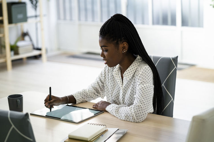 Woman writing on digital tablet while sitting in living room at home