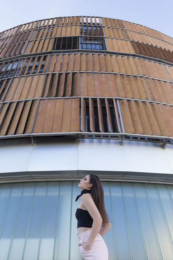 Fashionably dressed young woman looking at camera while standing near modern building in the city