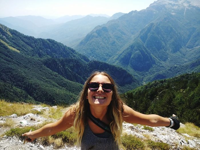 High angle portrait of cheerful young woman wearing sunglasses while standing against mountain ranges during sunny day