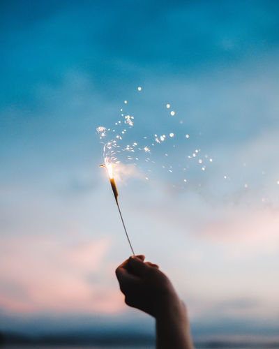 Cropped hand holding sparkler against sky during sunset