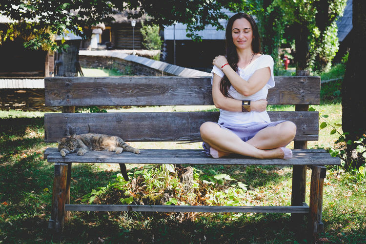 Young woman hugging self by cat on bench at park