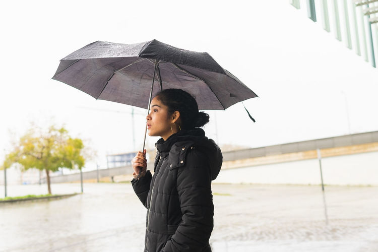 Side view of colombian female in black jacket standing with umbrella on street in rainy day and looking away with eyes wide open