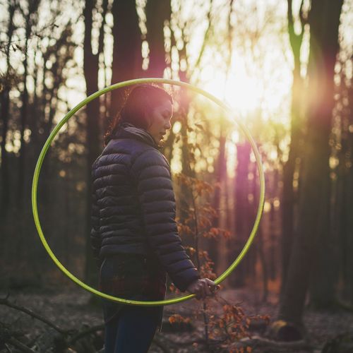 Side view of young woman holding plastic hoop in forest during sunset
