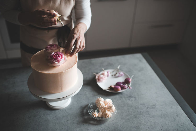 Woman making cake decorating with flower roses staying on kitchen table close up at home. 
