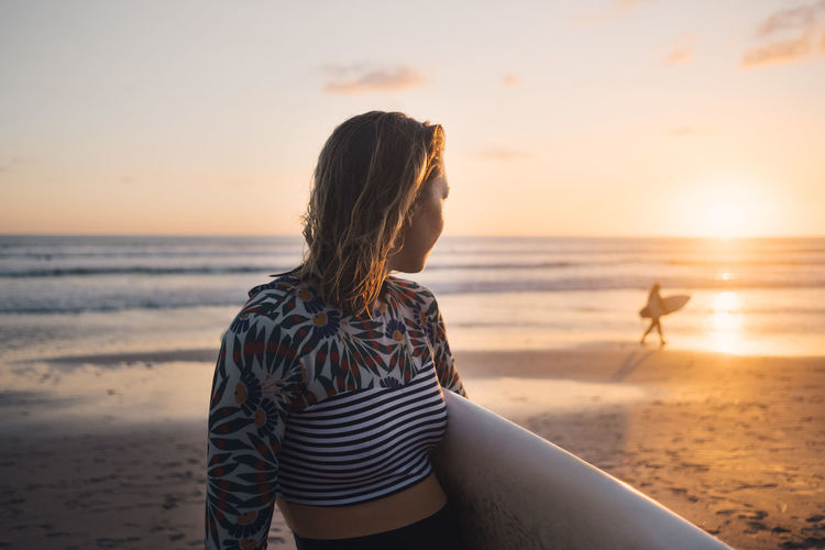 Mid adult woman with surfboard looking at sea during sunset at beach