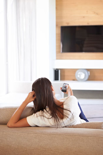 Rear view of woman switching on tv with remote control at home