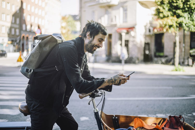 Side view of smiling businessman using smart phone while leaning on cargo bike