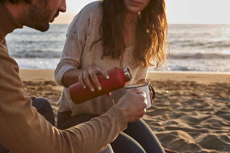 Woman pouring coffee in cup while sitting by man on beach