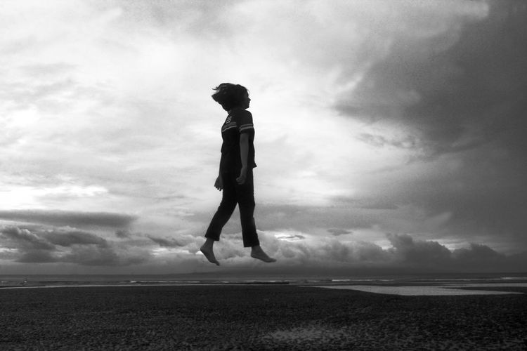 Side view of woman levitating at beach against cloudy sky