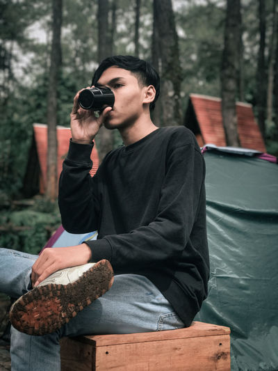 Young man holding camera while sitting on tree in forest