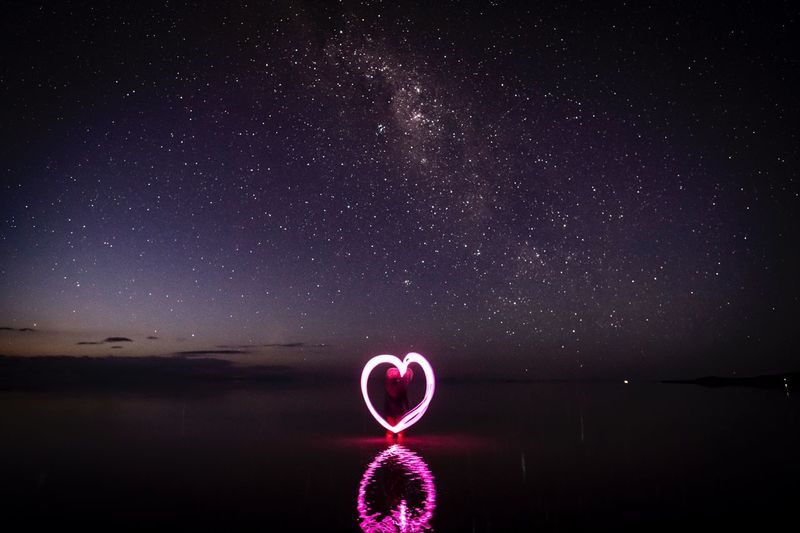 Woman making heart shape with light painting in sea against sky at night