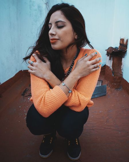 Full length of young woman with eyes closed hugging self while crouching against wall