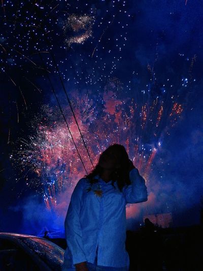 Woman standing by car against fireworks in sky at night