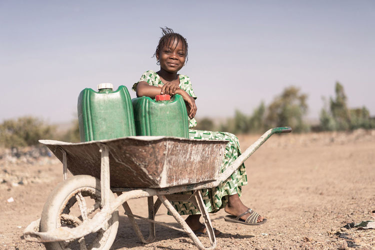 Full length portrait of girl sitting on wheelbarrow at land